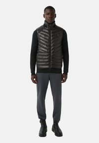 Zwart puffer-vest met een hoge kraag en ritssluiting, met een chevron-patroon, gecombineerd met donkergrijze broek en zwarte enkellaarsjes.