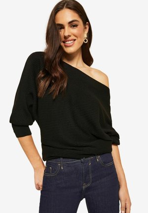 Sort ribbet strikket off-the-shoulder top med lange ærmer. Sættes sammen med mørkeblå jeans, der viser en afslappet pasform og draperet design.