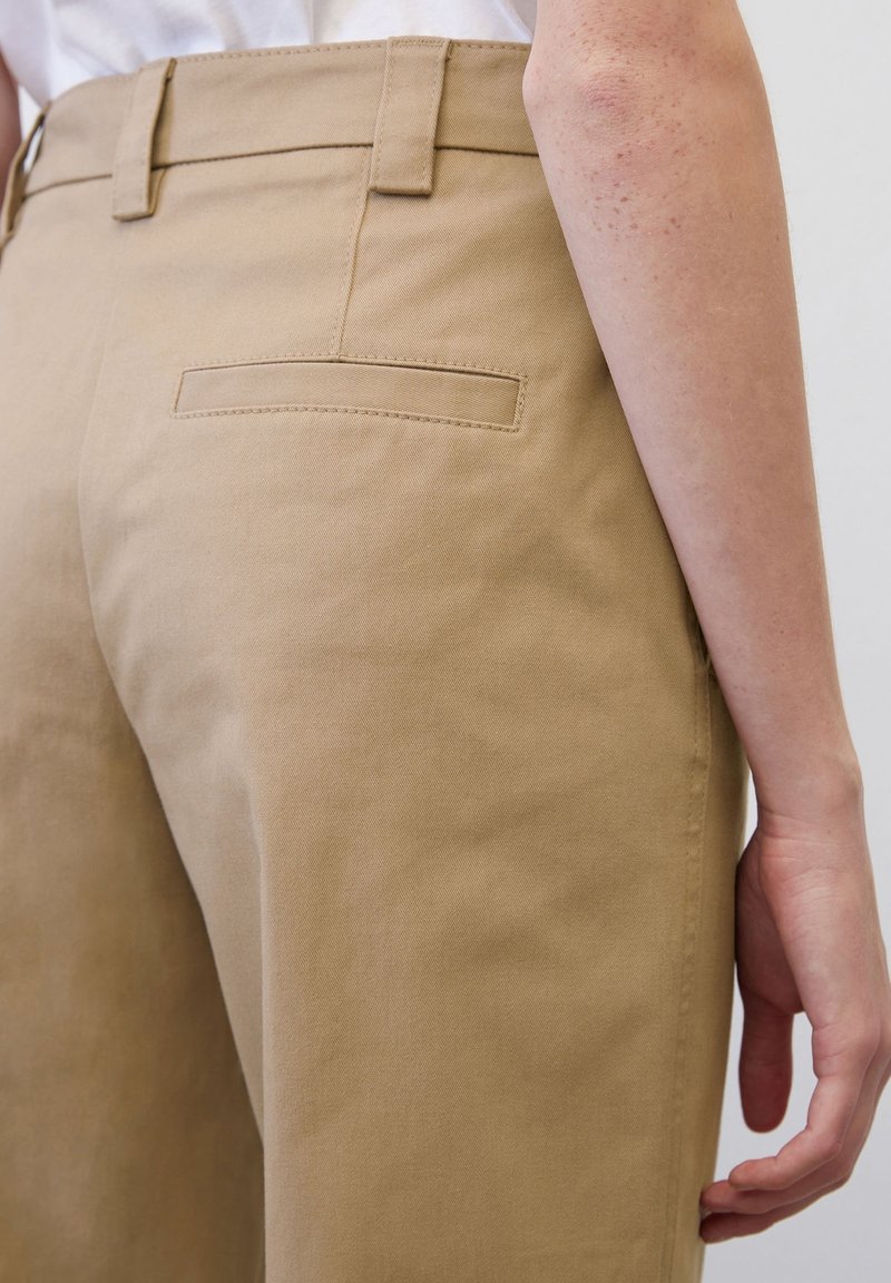 Gros plan sur une personne portant un pantalon beige taille haute avec passants pour ceinture et une poche arrière fendue, debout avec le bras le long du corps.