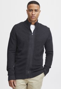 Črn zip-up pulover z rebrasto teksturo in visokim ovratnikom, narejen iz mehkega materiala. Nošen nad belo majico, v kombinaciji s svetlimi khaki hlačami.