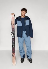 Tmavě modrá a světle modrá zateplená lyžařská bunda na zip, doplněná světle modrými lyžařskými kalhotami. Lyžař drží černo-béžové lyže s vázáním.