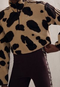 Fleece-pullover i beige med svart cowprint, med hög krage och halv dragkedja. Matcher med mörka leggings med mönstrade sidostråk.