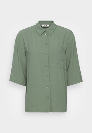 Chemise verte à manches courtes en tissu texturé, avec un col et une poche poitrine. Détails de couture discrets le long des boutons.