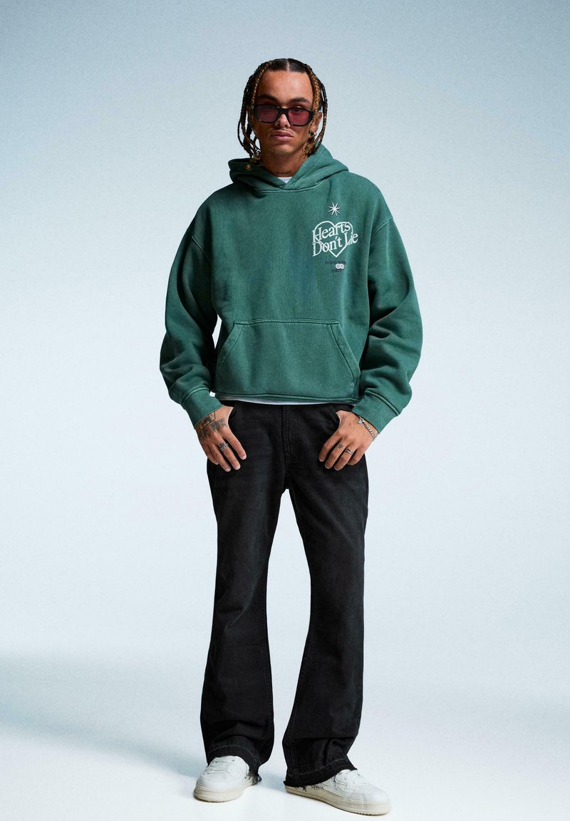 Homme aux cheveux tressés et lunettes de soleil, portant un sweat à capuche vert avec le texte "Hearts Don’t Lie", un pantalon noir et des baskets blanches, debout devant un fond uni.