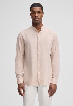 Man staat recht voor zich uit kijkend en draagt een lichtbeige overhemd met mandarin kraag en witte broek tegen een effen achtergrond.