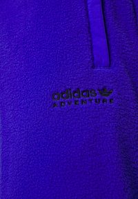 Lila fleecejacka med en kontrasterande blå blixtlås och svart "adidas ADVENTURE"-logga. Mjuk textur med en åtsittande design.