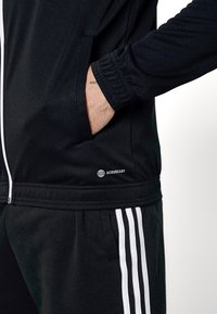 Schwarze Sportjacke mit weißem Reißverschluss und Aeroready-Logo; ausgestattet mit elastischen Bündchen und Seitentaschen. Kombiniert mit schwarzen Hosen und weißen Streifen.