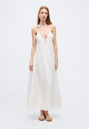 Femme portant une robe maxi blanche sans manches avec de fines bretelles et un décolleté en V profond, accompagnée de sandales tissées beiges à enfiler.