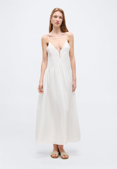 Woman wearing a sleeveless white maxi dress with thin straps and a deep V neckline, paired with beige woven slip-on sandals.
