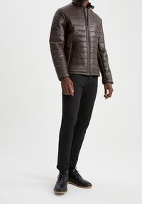 Giacca in pelle marrone trapuntata con collo alto, chiusura a zip e due tasche laterali, abbinata a pantaloni neri e scarpe nere.