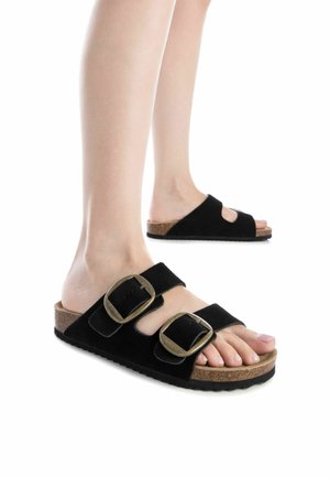 Sandalias de ante negro tipo slide con dos hebillas de bronce sobredimensionadas, plantilla de corcho y suela de goma texturizada. Diseño con los dedos y el talón al descubierto.