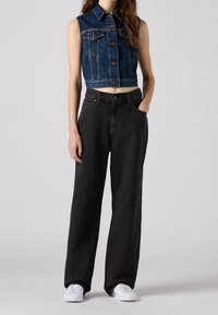 Denim crop vest in dark blue with front pockets and buttons, paired with high-waisted black wide-leg jeans and white sneakers.
