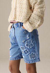 Shorts en denim bleu clair avec broderies florales, bords effilochés et taille haute. Portés avec un cardigan texturé crème.