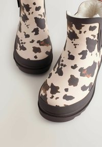 Brune og beige gummistøvler til anklerne med et cowprint-mønster. Struktureret sål og topkant, fuzzy foring synlig ved åbningen.