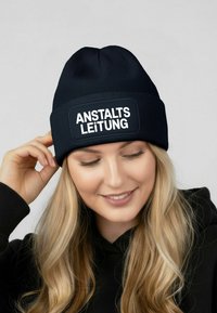 Navy knitted beanie with a rectangular black patch displaying the text "ANSTALTSLEITUNG" in bold white letters. Soft texture.