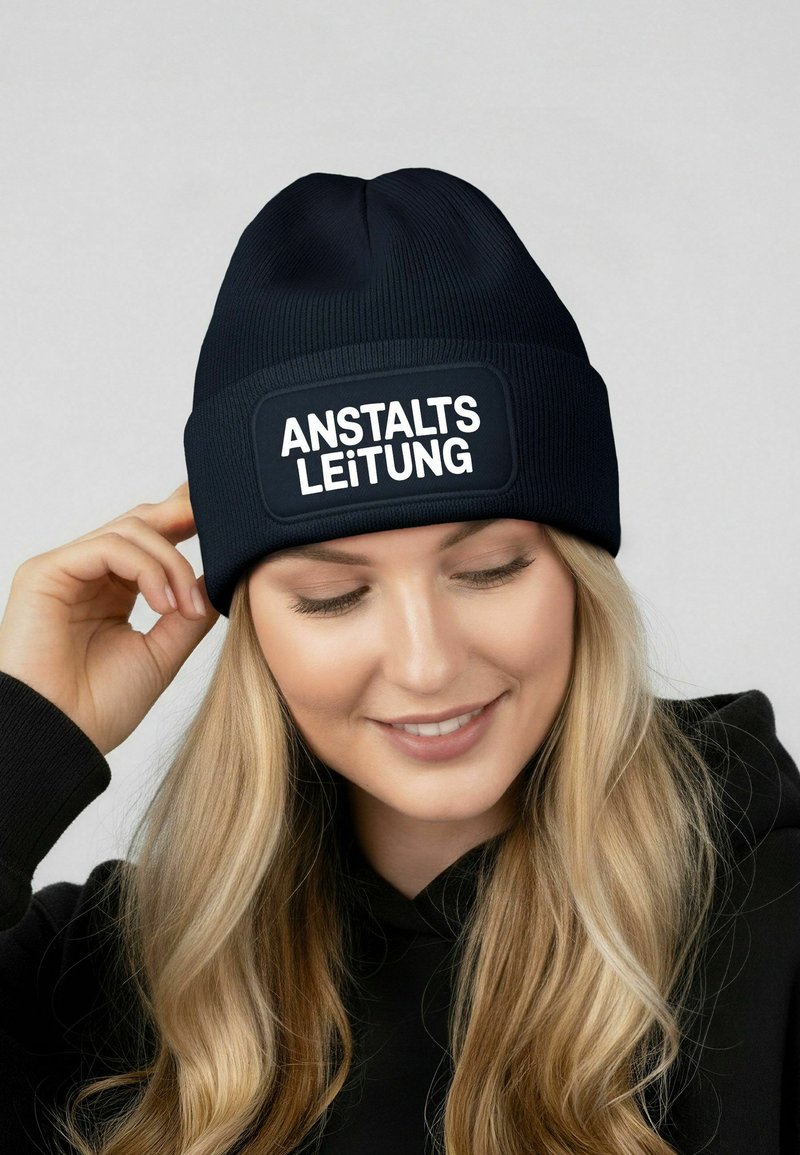 Navy knitted beanie with a rectangular black patch displaying the text "ANSTALTSLEITUNG" in bold white letters. Soft texture.