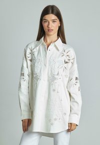 Camisa blanca de botones con intrincados bordados florales, corte holgado, mangas largas y un dobladillo ligeramente curvado para un aspecto moderno.