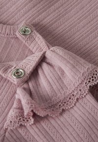 Lichtroze ribstof met een rimpelaccent en een zilveren drukknoopsluiting. De textuur is zacht met kantdetails aan de rand.