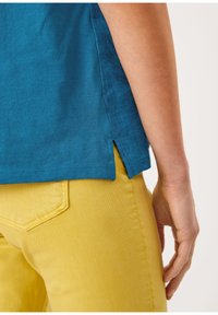 Primer plano de una persona con una camiseta de color azul turquesa con abertura lateral y pantalones amarillos, con la mano relajada a su lado frente a un fondo sencillo.