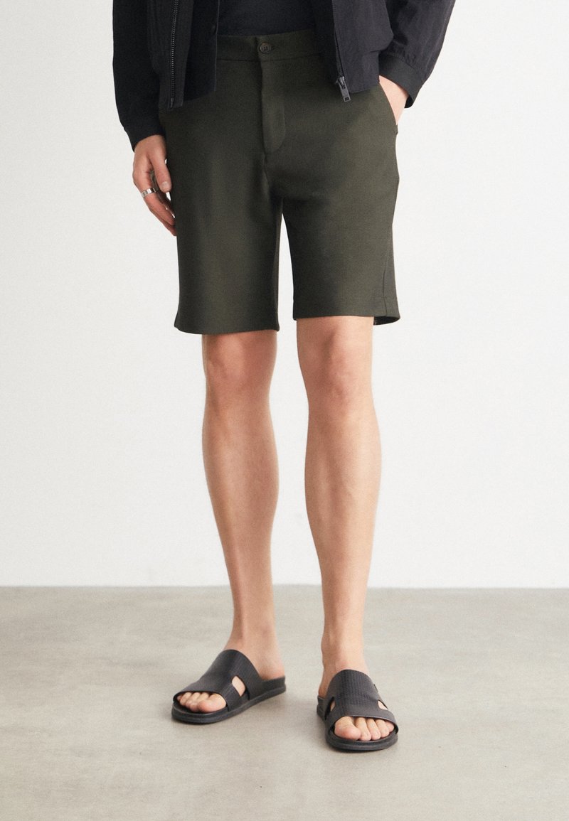 Personne portant un short vert foncé au genou, des sandales noires à enfiler et un haut noir à manches longues, les mains dans les poches.