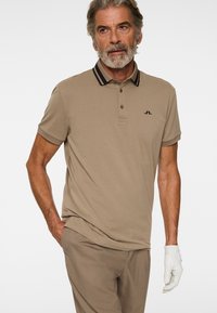 Starszy mężczyzna w beżowej koszulce polo z czarnym paskiem na kołnierzyku, pasujących spodniach oraz białej rękawiczce golfowej na prawej dłoni.