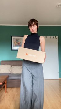 Kartono dėžė su oranžiniu Zalando logotipu, laikoma asmens, vilkinčio juodą be rankovių viršutinį drabužį ir pilkas plačias kelnes, gyvenamojoje erdvėje.
