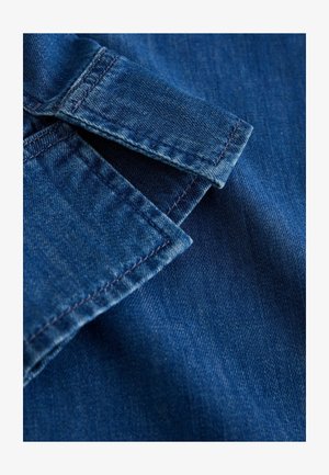 Džinsinis audinys su gilios mėlynos spalvos atspalviu, aiškiai matomu apykaklės kraštu ir siūlių detalėmis, lygiu paviršiumi ir švelniais blukimo raštais.