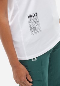 T-shirt en coton blanc avec un imprimé graphique noir étiqueté "MILLET" avec un motif floral. Associé à un pantalon cargo vert foncé présentant un logo.