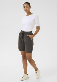 Shorts en coton vert olive foncé avec une taille à cordon, poches avant, poignets retroussés, associés à une chemise blanche à manches courtes et des baskets beiges.