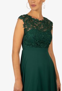 Grünes Kleid mit floralem Spitzenoverlay auf einem taillierten Oberteil, dazu ein sanft fließender Rock. Kurze Ärmel und runder Ausschnitt.