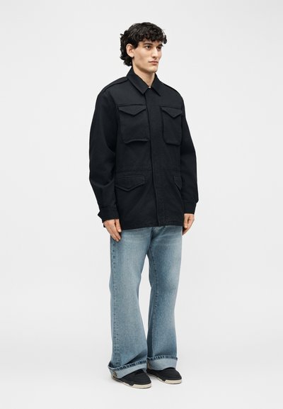 Νέος άνδρας με σκούρα σγουρά μαλλιά, φορώντας μαύρο σακάκι με κουμπιά και τέσσερις μπροστινές τσέπες, ανοιχτόχρωμα φαρδιά τζιν παντελόνια και μαύρα αθλητικά παπούτσια.