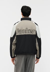 Άνδρας που φοράει μπουφάν με σχέδιο χρωμάτων και τη λέξη «Jacker» κεντημένη στην πλάτη, βλεπόμενος από πίσω με φόντο ένα απλό τοπίο.