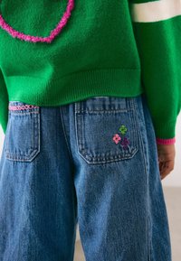 Vihreä neulepaita, jossa on vaaleanpunaista yksityiskohtaa ja sydänkuvio, yhdistettynä sinisiin denim-farkkuihin, joissa on brodeerattuja kukkia takataskuissa.