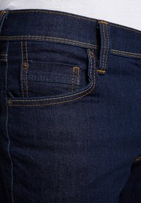 Tumedast sinisest denimpüksist, millel on oranž õmblus, esiküljel tasku ja metallist nööbiga detail vöökohtadel. Sile tekstuur.