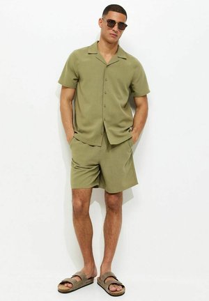 Homme portant une chemise à manches courtes boutonnée vert olive et un short assorti, des lunettes de soleil et des sandales marron, debout les mains dans les poches.