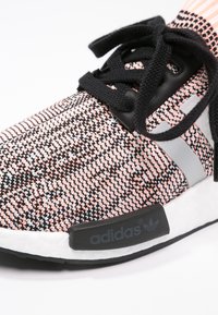 Close-up of a knit Adidas sneaker in pink, black, and white with black laces and signature side stripes on a white sole.