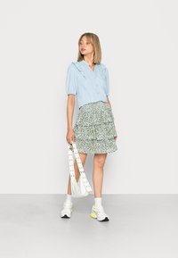 Camisa azul claro com botões e detalhes franzidos, combinada com uma saia verde com camadas em estampado floral. Bolsa branca com tachas e ténis brancos.