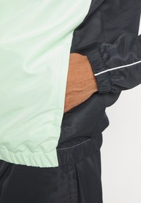 Šviesiai žalias ir juodas vėjo jaku, pagamintas iš tekstūruoto audinio, su elastingomis rankovėmis, užtrauktuku užsegama šonine kišene ir kontrastinga balta siūle.