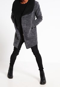 Abrigo gris de mezcla de lana con un gran cuello negro, cremallera frontal y bolsillos laterales, combinado con jeans ajustados negros y botas con cordones.