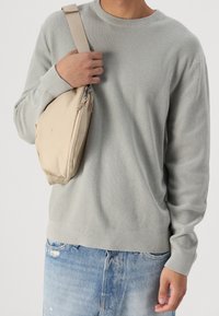 Persoon in een lichtgrijze gestructureerde trui en lichtblauwe ripped jeans, met een beige schoudertas over de schouder, tegen een witte achtergrond.