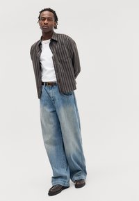 Braun gestreifte Hemd, weißes T-Shirt, hellblaue weit geschnittene Jeans, dunkle Schuhe, stehende Pose, neutraler Hintergrund.