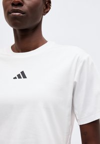 Κοντινό πλάνο ατόμου που φοράει λευκό t-shirt Adidas με μαύρο λογότυπο στο στήθος και πολλαπλά χρυσά σκουλαρίκια στο αριστερό αυτί.