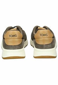 Šedé semišové tenisky s hnědým koženým akcentem na patě, bílé mezipodešvi a hnědou podešví. Na zadní straně je logo "TOMS".