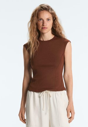 Žena s vlnitými blond vlasmi, ktorá má na sebe hnedý priliehavý top bez rukávov a krémové nohavice so šnúrkou, stojaca pred jednoduchým svetlým pozadím.