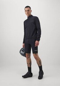 Svart cykeljacka med hög krage, kombinerad med svarta vadderade shorts. Svarta cykelskor och en grå hjälm. Synliga tatueringar på benen.