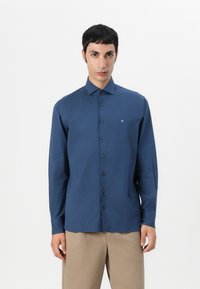 Chemise à manches longues bleu marine avec un col boutonné, présentant un logo discret sur la poitrine et une texture lisse et mate.