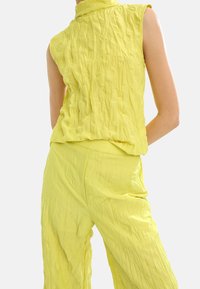 Limonkowy, bezrękawnik z wysokim kołnierzem, wykonany z teksturowanej, pomarszczonej tkaniny. W połączeniu z dopasowanymi, luźnymi spodniami.