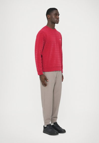 Κόκκινο φούτερ με υφή και γεωμετρικό μοτίβο, συνδυασμένο με ανοιχτό γκρι παντελόνι με στενή γραμμή και μαύρα αθλητικά παπούτσια. Διαθέτει μικρό λογότυπο στο στήθος.