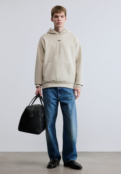 Sweat à capuche beige avec une poche avant, porté avec un jean ample bleu clair. Sac de voyage noir de forme arrondie avec de courtes poignées. Chaussures noires.
