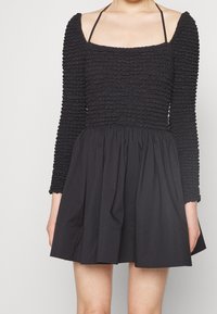 Svart klänning med en texturerad, volangad överdel, långa ärmar, fyrkantig halsringning och en utsvängd kjol. Materialet verkar vara lätt.